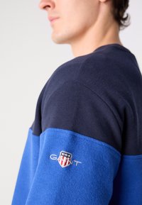 Blue and navy sweater with ribbed texture, featuring an embroidered logo on the sleeve. The design includes color-blocking across the chest.