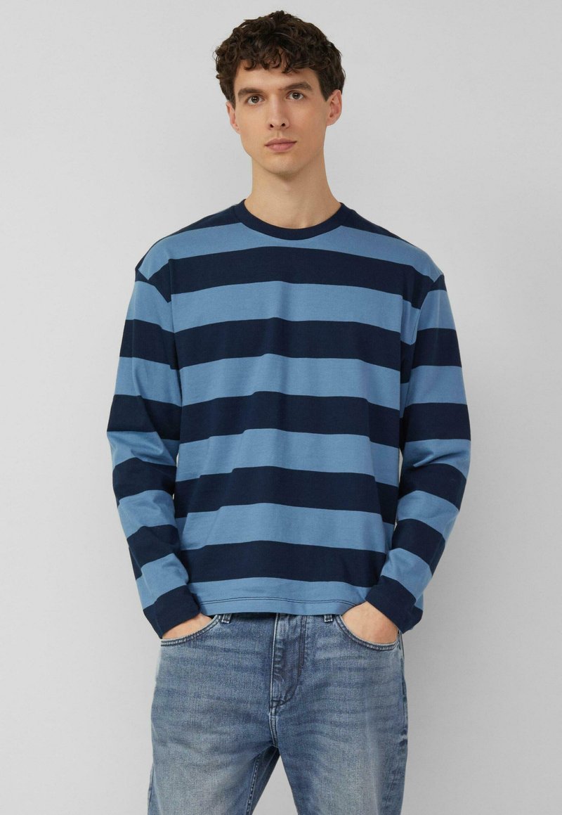 Mladić obučen u plavu i tamnoplavu vodoravno prugastu majicu s dugim rukavima i svijetloplave traperice s rukama u džepovima, stoji naslonjen na jednoličnu pozadinu.