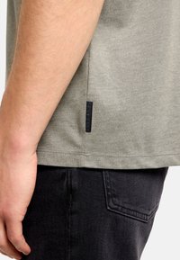 Sivé bavlnené tričko s postrannou značkou a krátkymi rukávmi, v kombinácii s čiernymi rifľovými nohavicami. Textília pôsobí mäkko a v neformálnom štýle.