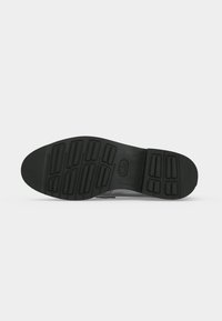 Semelle d'une chaussure noire avec des motifs de crampons rectangulaires texturés pour une bonne adhérence, présentée sur un fond gris clair uni.