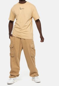 Beige puuvillainen t-paita, jossa musta skriptilogolla, yhdistetty beigenvärisiin corduroy-housuihin, joissa on sivutaskut. Valkoiset tennarit viimeistelevät asun.