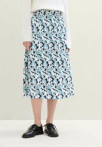 Frau trägt einen blauen, weißen und schwarzen blumenmusternden plissierten Midirock mit einem weißen Pullover und schwarzen, schnürbaren Lederschuhen.