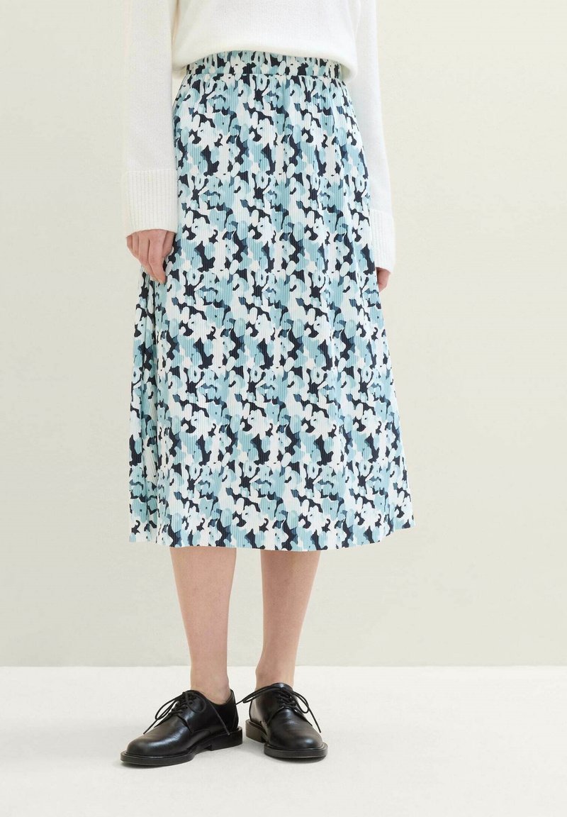 Frau trägt einen blauen, weißen und schwarzen blumenmusternden plissierten Midirock mit einem weißen Pullover und schwarzen, schnürbaren Lederschuhen.