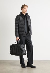 Giacca a vento nera con texture trapuntata, maglietta nera, pantaloni e scarpe nere. Porta una grande borsa da viaggio in pelle nera con doppi manici.