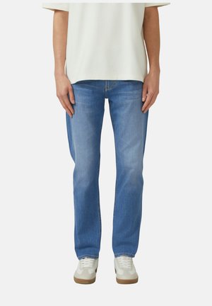 Person, die hellblaue Jeans mit geradem Bein, weiße Sneakers und ein cremeweißes kurzärmeliges Oberteil trägt, steht mit locker an den Seiten hängenden Armen.