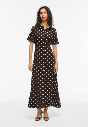 Kobieta stoi, ubrana w czarną długą sukienkę z krótkim rękawem w białe kropki i z guzikami, połączoną z brązowymi, otwartymi na palce szpilkami.