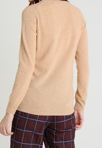 Osoba w beżowym swetrze z długim rękawem i brązowych spodniach w kratę, pokazana od tyłu na tle jasnym.