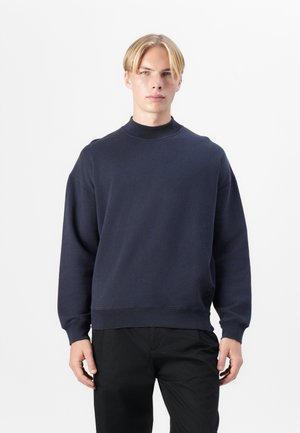 Pulover navy cu guler înalt, croială supradimensionată, manșete și tiv cu nervuri. Fabricat dintr-un material moale, texturat, cu un accent subtil al logo-ului.