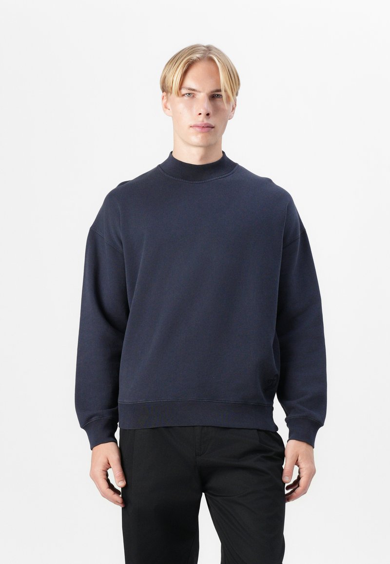 Sötétkék pulóver magas nyakkal, oversize fazonnal, bordás mandzsettákkal és szegéllyel. Puha, texturált anyagból készült, finom logóval díszítve.