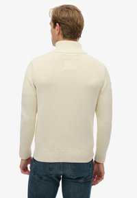 Cremefarbener Rollkragenpullover mit gerippten Bündchen und Saum, gestrickte Textur, figurbetonte Silhouette, von hinten gezeigt, kombiniert mit blauer Jeans.