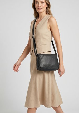 Kvinde iført beige ærmeløs kjole med sort læder crossbody taske, stående mod en ensfarvet lys baggrund.