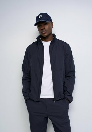 Man wearing a navy blue jacket, white t-shirt, matching navy pants, and a navy cap with a "SALSA JEANS" logo standing against a plain white background.
