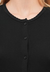 Cardigan negru cu dungi, cu decolteu în dantelă și cinci nasturi negri. Țesătura pare texturată și ajustată în zona gulerului.