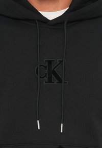 Sweatshirt preta com capuz feita de tecido macio. Apresenta o logótipo "CK" bordado a preto, cordões ajustáveis com pontas metálicas e um bolso canguru.