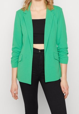 Femme portant un blazer vert vif par-dessus un crop top noir et un pantalon noir taille haute sur un fond blanc uni.