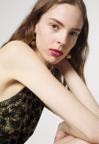 Close-up de uma modelo com cabelo longo, vestindo um vestido verde sem mangas com um padrão de folhas pretas, e brincos dourados com esferas texturizadas.