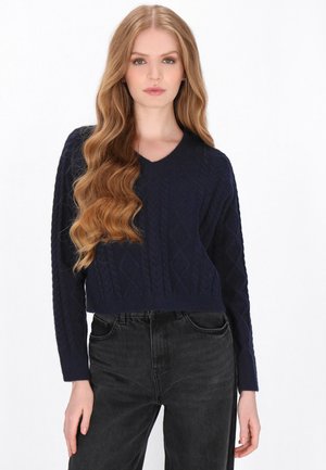 Ung kvinde med langt, bølget, blondt hår iført en mørkeblå kabelstriktrøje og sorte højtaljede jeans mod en hvid baggrund.