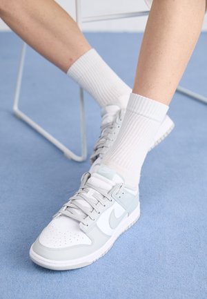 Nogi w białych prążkowanych skarpetkach i szaro-białych butach Nike odpoczywające na jasnoniebieskim dywanie, z widoczną częścią białego metalowego krzesła.