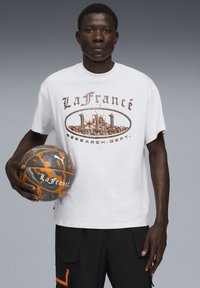 Hvid t-shirt med korte ærmer, der har et "LaFrance" grafisk print i orange og brun. Modellen holder en sort og orange mønstret basketball. Sorte cargo shorts.