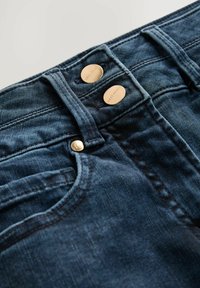 Jeans de mezclilla de un azul oscuro con un diseño clásico de cinco bolsillos, que cuentan con botones de metal dorados y costuras en contraste. Textura suave, ajuste a medida.