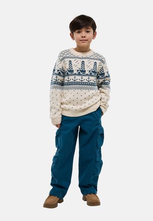 Jeune garçon debout, les mains dans les poches de son pantalon cargo bleu, portant un pull beige avec un motif de phare bleu et d’ancre, des chaussures marron.