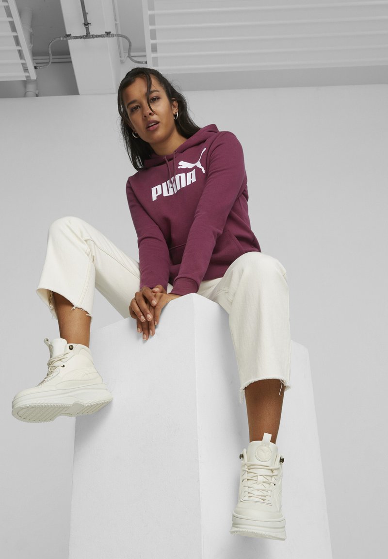 Maroon hoodie with white Puma logo, cream wide-leg pants with frayed hem, and white high-top sneakers, positioned on a white pedestal.