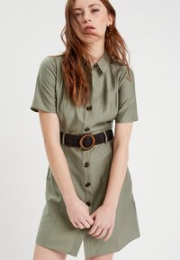 Khakifarbenes Hemdkleid mit kurzen Ärmeln, durchgehender Knopfleiste und einer Taille, die durch einen schwarzen geflochtenen Gürtel mit einer runden Holzschnalle betont wird.
