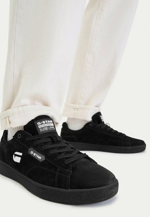 Person wearing white pants and black G-Star low-top sneakers with lace-up design and branded labels on tongue and side.