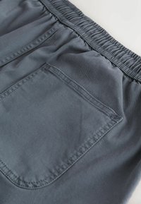 Gros plan sur un pantalon à taille élastique grise montrant une poche arrière cousue avec des détails de coutures visibles.