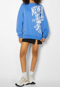 Osoba oblečená v modrém oversized svetru s bílým grafickým textem, černé mini sukni, bílých ponožkách a pastelově barevných chunky teniskách.