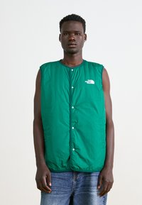 Green insulated vest with a high collar, snap buttons, and logo on the left chest. Worn over a dark top and light denim jeans.
