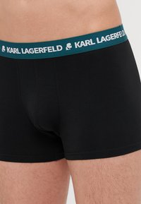 Melnas bokseru bikses ar zilu-zaļu elastīgu jostu, kurā ir balta "KARL LAGERFELD" logo. Gluda tekstūra, pieguļoša forma.