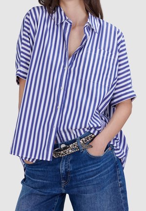 Frau, die ein lockeres blau-weiß gestreiftes Kurzarmshirt, blaue Jeans und einen genagelten schwarzen Gürtel trägt, mit den Händen in den Taschen.