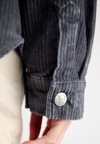 Sivá kordurová manžeta košele s vertikálnou ryhovanou textúrou, so strieborným označeným gombíkom s logom srdca. Ruka viditeľná v zábere.