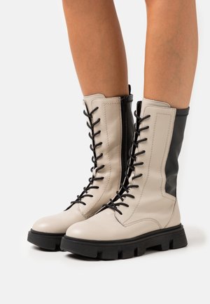 Bottes de combat à lacets crème et noires avec semelles épaisses noires portées sur des jambes nues sur un fond blanc.