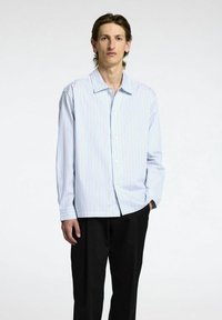 Skjorta med knappar i ljusblått med vertikala ränder i gult och rosa. Långa ärmar med enstaka knappar vid ärmsluten. Bärs med svarta byxor.