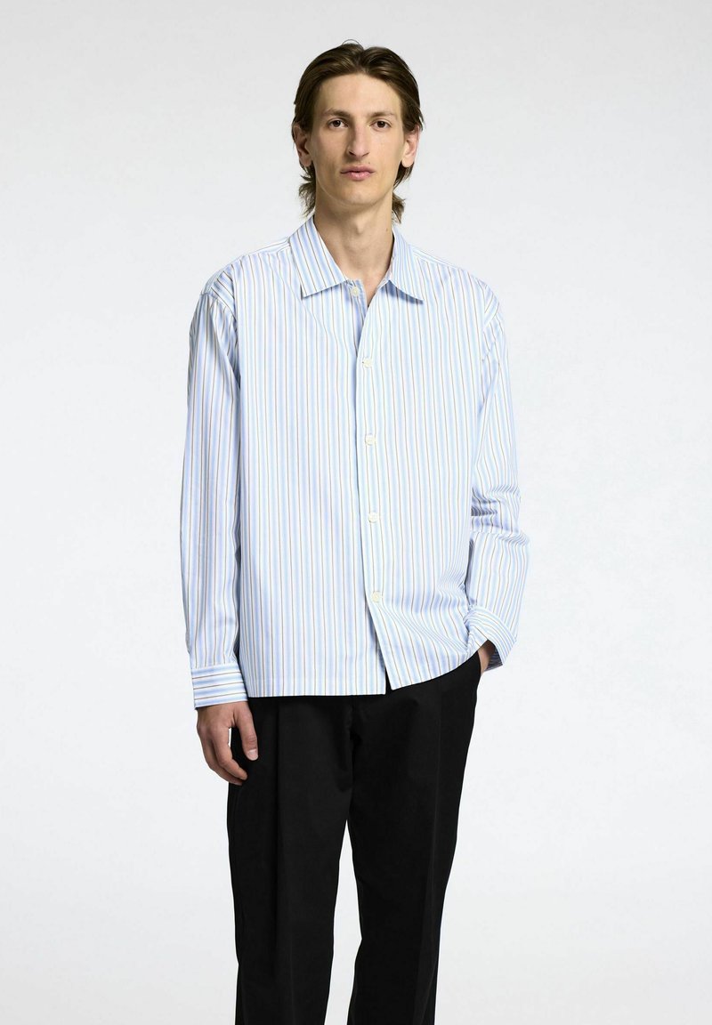 Skjorta med knappar i ljusblått med vertikala ränder i gult och rosa. Långa ärmar med enstaka knappar vid ärmsluten. Bärs med svarta byxor.