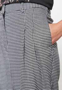 Checkered black and white trousers with a houndstooth pattern, featuring a loose fit, pleats, and side pockets. Fabric appears textured.