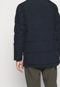 Marinblå pufferjacka med kviltad design, inklusive långa ärmar och rak nederkant. Mjuk, matt textur utan synliga knäppningar eller utsmyckningar.