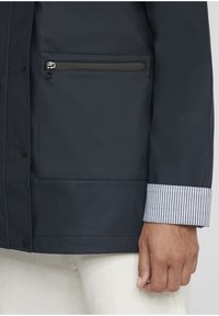 Veste imperméable marine avec une poche zippée sur la poitrine, des détails de poignet rayés et des boutons-pression à l'avant, mettant en valeur une finition lisse et texturée.