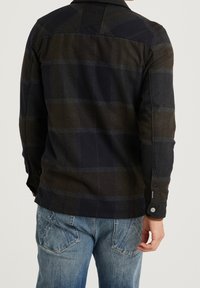 Muž v tmavé kárované bundě s zapnutými manžetami a modrých džínách, zobrazený zezadu na jednobarevném pozadí.