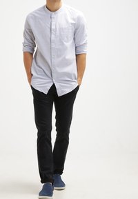 Hombre con una camisa de botones gris claro y mangas remangadas, pantalones negros y zapatos azul marino sin cordones, de pie con las manos en los bolsillos.