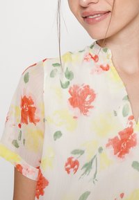 Květinová blůza vyrobená z lehké, průhledné látky. Disponuje červenými, žlutými a zelenými květinovými vzory, výstřihem do V a krátkými, mírně nabíranými rukávy.