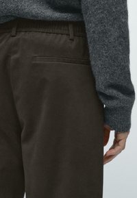 Pantaloni marroni in materiale morbido, con vita elastica e tasca posteriore, abbinati a un maglione grigio testurizzato.