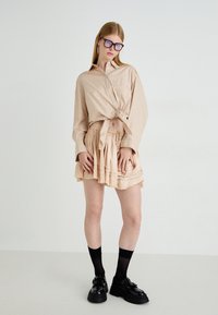 Lichtbruine geruite overhemdo geknoopt in de taille, gecombineerd met een geplooide beige rok. Model draagt zwarte chunky schoenen en kniehoge sokken.