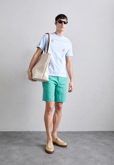 Ljust blå t-shirt med broderi, gröna shorts, beige tote bag och vävda loafers, med en avslappnad design och neutral färgpalett.