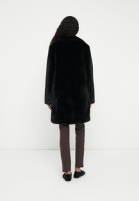 Person med langt krøllet hår iført en sort faux fur frakke, mørkebrune slim pants og sorte strukturerede loafers, stående foran en hvid baggrund.