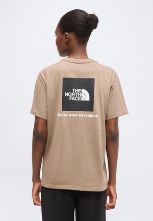 Personne à la peau foncée et aux cheveux attachés en chignon portant un t-shirt beige avec le logo The North Face et le texte "Never Stop Exploring" au dos.