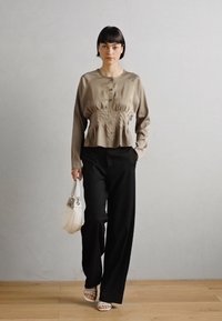 Blouse beige à manches longues avec boutons et taille froncée, associée à un pantalon large noir. Le modèle porte un sac blanc et des sandales blanches.