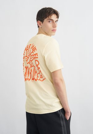 Jeune homme aux cheveux courts et foncés portant un t-shirt crème avec un grand papillon orange et du texte au dos, les mains dans les poches de son pantalon noir.